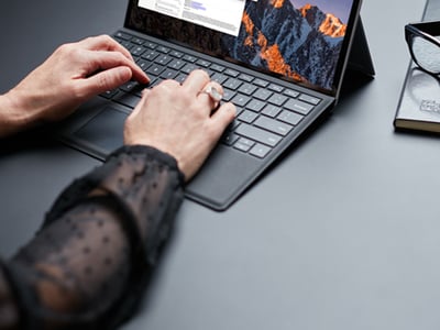 Person typing on a tablet with keyboard cover, displaying code and a mountain landscape wallpaper, with a Six Degrees branded notebook and glasses nearby on a dark desk.
