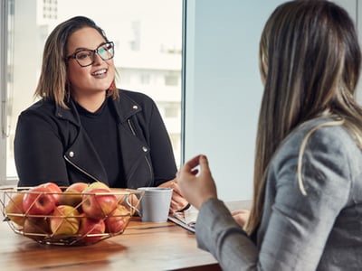 Two women having a professional conversation at a wooden table with a bowl of apples, in a bright office setting.