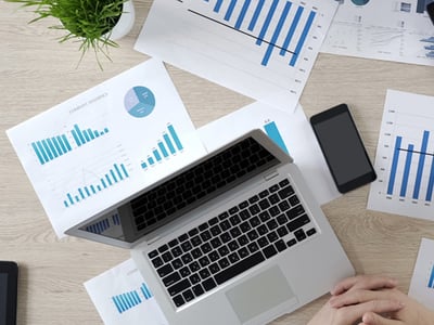 Top view of a desk with a laptop, smartphone, and multiple printed charts and graphs showing business data in blue tones.