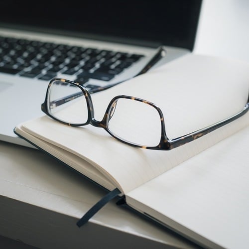 A pair of eyeglasses resting on an open notebook beside a laptop on a white desk.