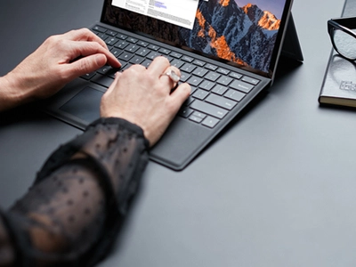 Person typing on a tablet with keyboard cover, displaying code and a mountain landscape wallpaper, with a Six Degrees branded notebook and glasses nearby on a dark desk.